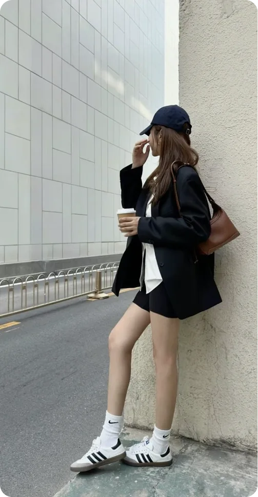 A woman leaning on a wall in a navy cap and navy blazer over a white top, with black shorts, a brown shoulder bag and white trainers