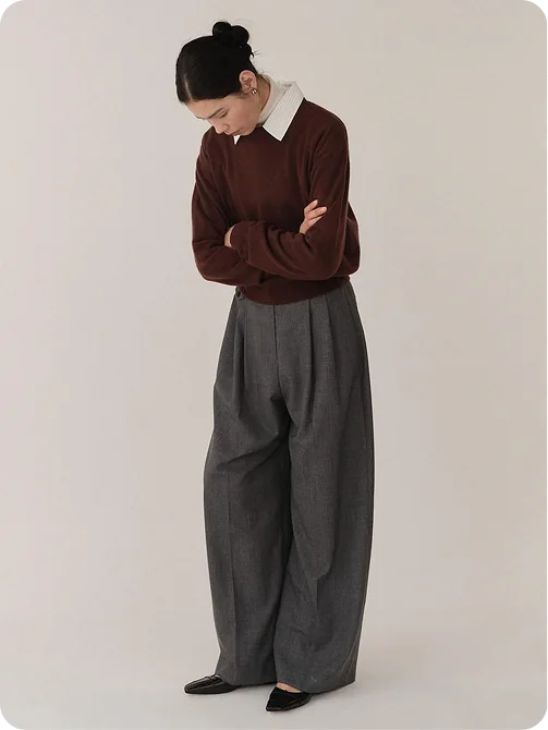 A woman in a burgundy jumper over a white collared shirt, with grey pleated wide-leg trousers and black flats