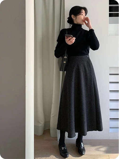A woman in a black turtleneck with a charcoal wool A-line midi skirt, black tights, loafers and a small black shoulder bag